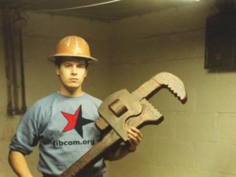 A man in a libcom t-shirt wearing a hard hat and carrying wrench nearly as big as he is