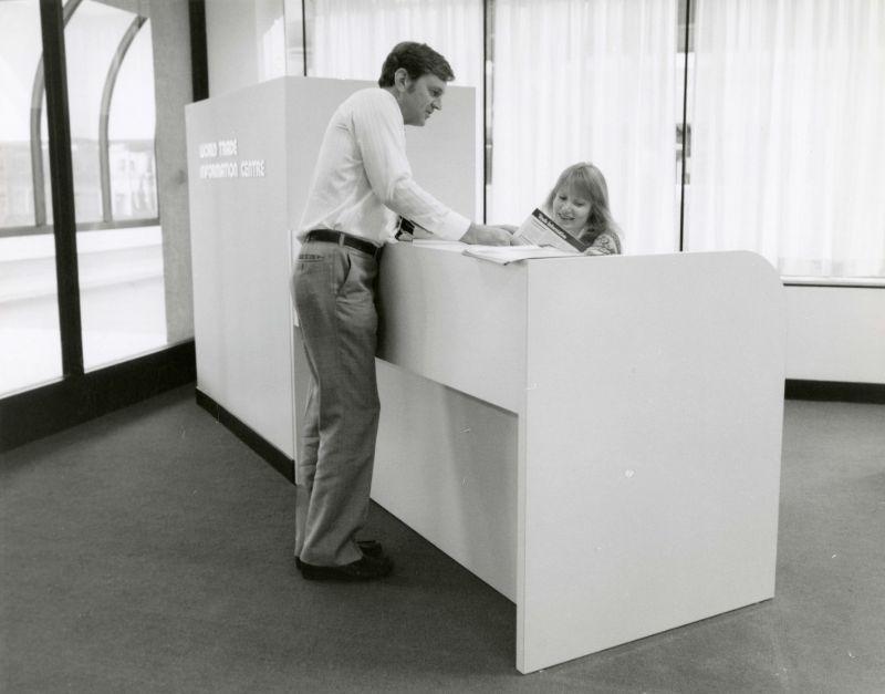 A man in a suit stands over a female receptionist in an office.